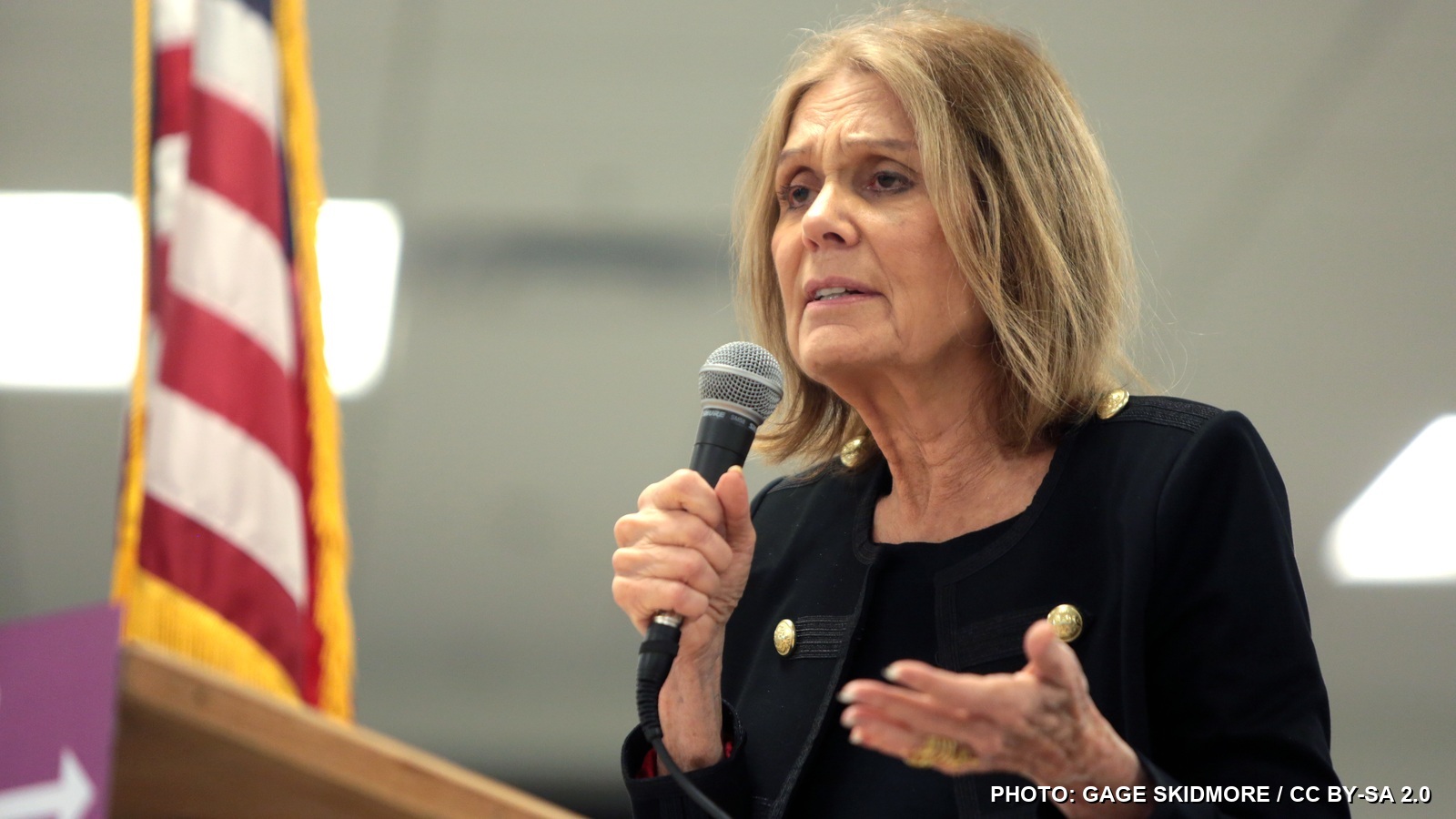 Gloria Steinem speaking into a microphone in 2016 with an American flag behind her